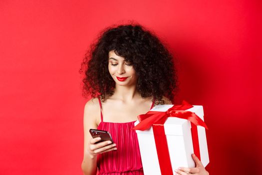 Valentines day and shopping. Beautiful caucasian woman in red dress, holding gift box from lover and using mobile phone, reading message on smartphone, studio background.