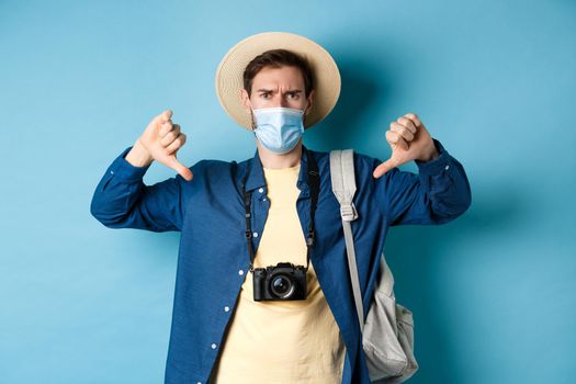 Covid-19 and summer holidays concept. Disappointed tourist in medicla mask frowning, showing thumbs down, negative feedback about travel, standing on blue background.