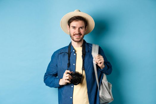 Cheerful handsome guy going on vacation, wearing summer hat and holding backpack with camera for photos, smiling excited of holiday, standing on blue background.