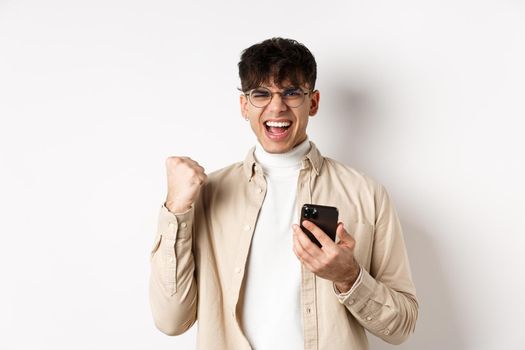 Technology and online shopping concept. Motivated and happy guy scream yes and show fist pump after winning on smartphone, achieve goal in app, white background.