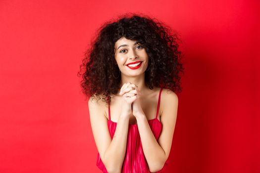 Image of beautiful curly-haired woman in red dress and makeup, saying thank you, looking grateful and smiling happy at camera, studio background.