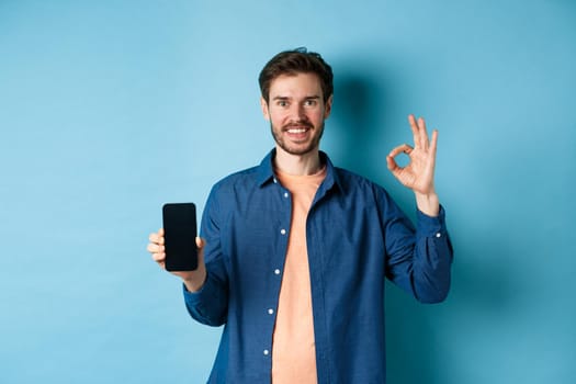 Handsome young man showing okay gesture and empty smartphone screen, recommending app or company, standing on blue background.