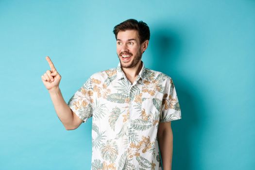 Excited smiling tourist pointing and looking aside at empty space, checking out promo offer, standing in hawaiian shirt on blue background.