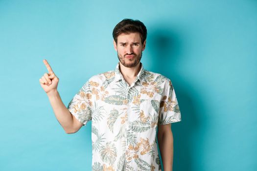 Disappointed tourist cringe and frown, pointing finger aside at empty space, complaining on bad thing, standing in hawaiian shirt on blue background.