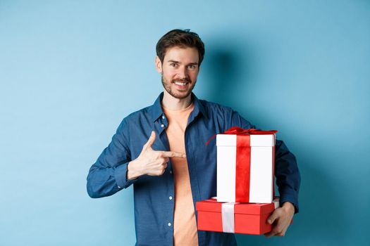 Attractive boyfriend pointing finger at valentines day surprise gifts, showing romantic presents for girlfriend, standing against blue background.