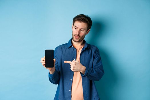 Image of handsome man talking about application, pointing finger at mobile screen and standing on blue background.