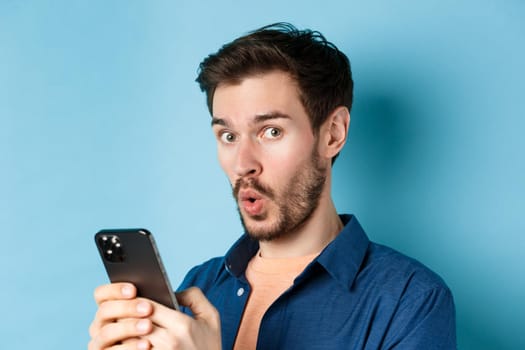 Close up of surprised caucasian man say wow, looking amused at camera, holding smartphone, standing on blue background.