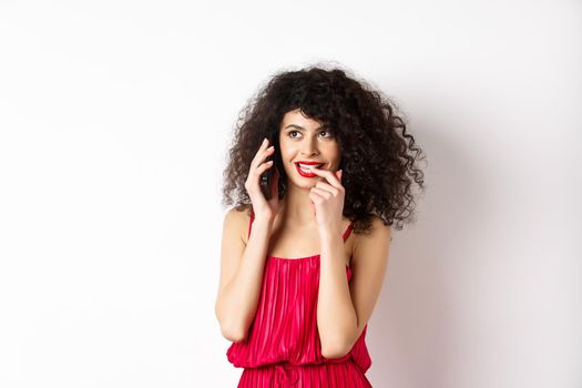 Silly fashionable woman in red dress and lips, biting fingernail duing phone call, thinking on mobile conversation, standing on white background.