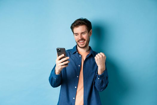 Image of lucky guy winning online, reading mobile phone screen and making fist pump, triumphing, standing on blue background.