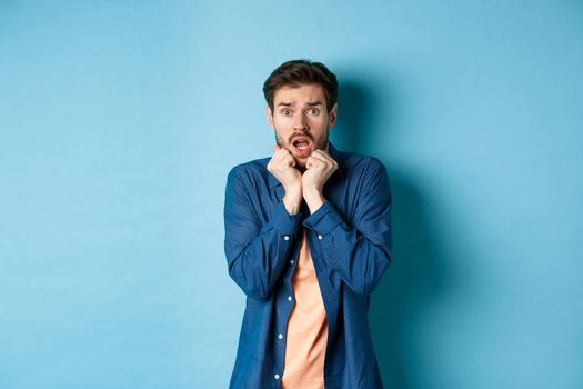 Scared young man gasping and screaming frightened, looking terrified at something, standing on blue background.