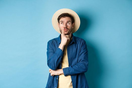 Thoughtful male tourist thinking of summer holiday vacation, looking aside with pensive face, touching lip and pondering next travel, standing on blue background.