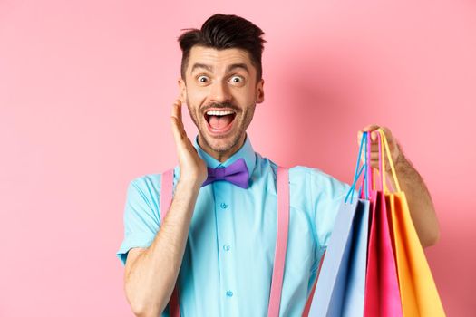 Surprised and happy man screaming of joy and holding shopping bags, shopper seeing discounts, standing over pink background.