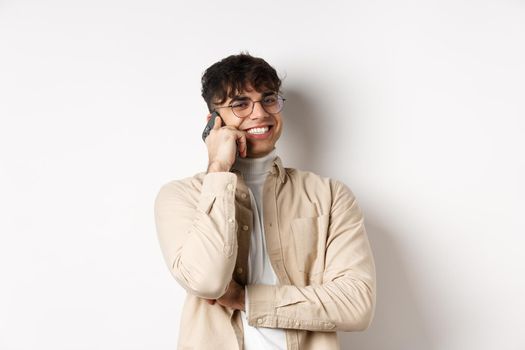 Real people concept. Happy young guy talking on mobile phone, look aside and holding smartphone near ear, standing on white background.