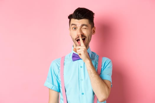 Funny young man planning a surprise, hushing and winking at camera, asking to keep quiet, telling a secret, standing on pink background.