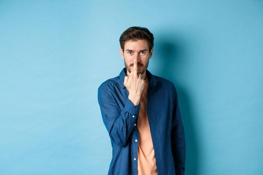 Ignorant and rude guy showing middle finger and smiling, say fuck you, standing on blue background.