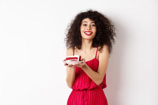 Beautiful lady in red dress celebrating birthday, holding piece of cake with candle and smiling, standing happy on white background.
