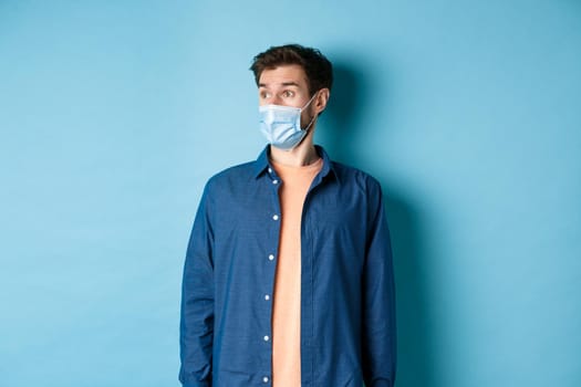 Covid-19 and healthcare concept. Surprised caucasian guy in face mask looking aside at empty space for banner, standing on blue background.
