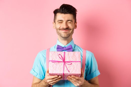 Holidays and celebration concept. Happy guy with french moustache grinning from delight, showing cute gift box, receiving present, standing over pink background.