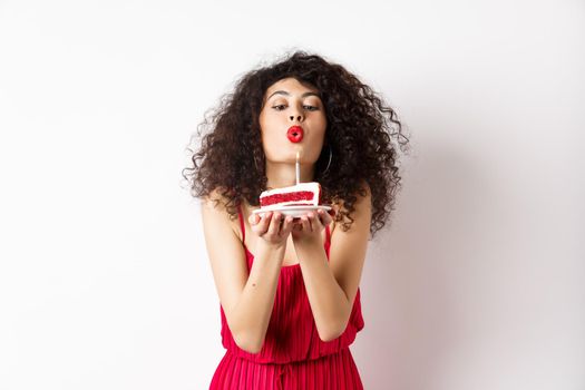 Holidays and celebration concept. Romantic woman in red dress celebrating birthday, blowing out candle on b-day cake and making wish, standing over white background.