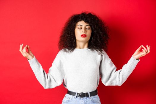 Woman in casual clothes trying to calm down with meditation, close eyes and practice yoga, holding hands sideways with zen gesture, standing on red background.