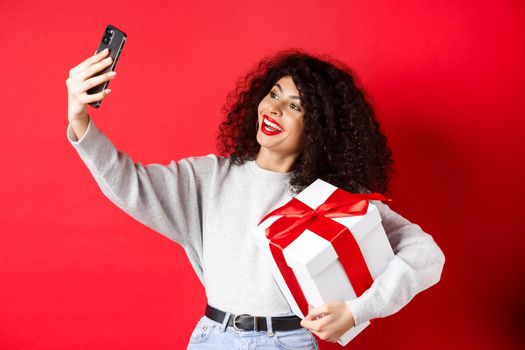 Holidays and tech concept. Happy woman taking selfie with her gift, holding present and smartphone, standing on red background.