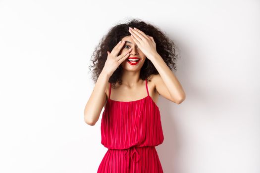 Surprise. Excited pretty girl with curly hair and red lips, peek through fingers and smiling amazed, checking out promo, standing on white background.