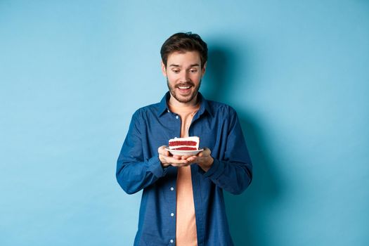 Celebration and holiday concept. Happy young man looking at birthday cake and making wish, standing on blue background.