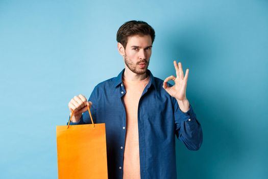 Handsome young man recommending store, showing orange shopping bag and OK sign, standing on blue background.