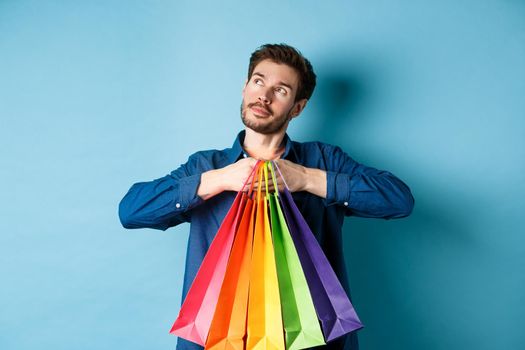 Dreamy young man holding colorful shopping bags and imaging something, looking at upper left corner empty space, blue background.