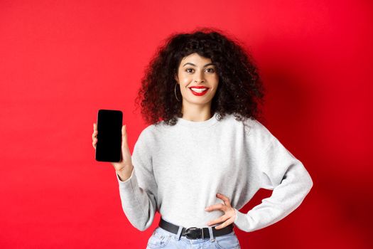 Attractive smiling woman showing empty smartphone screen and looking happy, advertising online store or application, standing against red background.