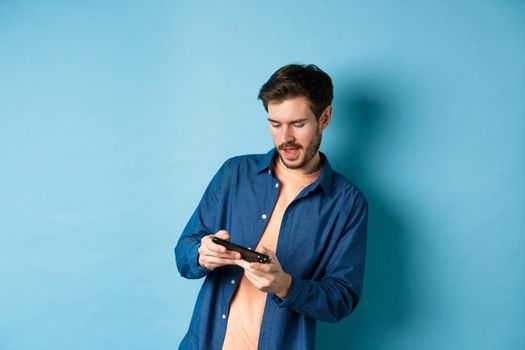 Young guy playing video game on mobile phone, tilt body and holding smartphone horizontally, standing on blue background.