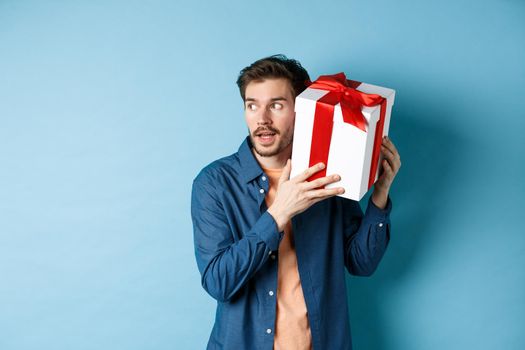 Valentines day. Curious boyfriend holding gift box near ear, trying to guess whats inside surprise present, standing over blue background.