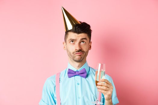 Holidays and celebration concept. Grumpy guy wearing birthday party hat and holding glass of champagne, looking up with skeptical face, standing on pink background.