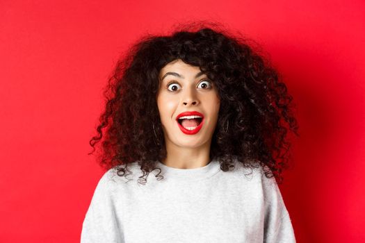 Close-up portrait of excited woman with curly hair, scream surprised and amazed, checking out special deal, standing on red background.