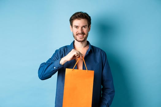 Handsome young man with beard, smiling and showing shopping bag, buying something in store, standing on blue background.