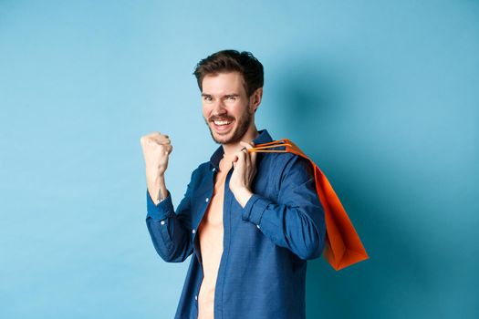 Cheerful guy saying yes, raising fist pump and smiling, holding orange shopping bag, feeling joy after buying with discounts, standing on blue background.