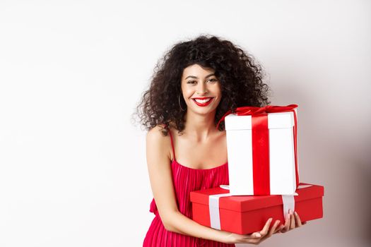 Beautiful birthday girl with curly hair, holding bday gifts and smiling happy, celebrating, standing against white background.