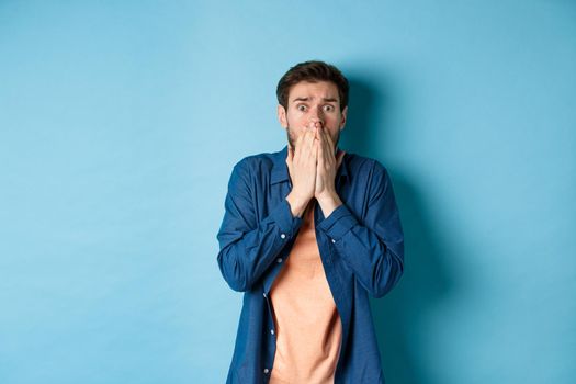 Scared anxious man covering mouth with hands and looking at something terrifying, standing on blue background.