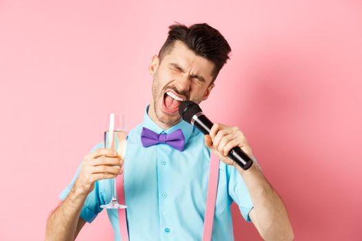 Party and festive events concept. Drunk funny guy singing in microphone and drinking champagne from glass, having fun at karaoke bar, standing on pink background.