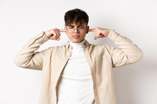 Annoyed young man shut his ears, block annoying sound, unwilling to listen, standing on white background.
