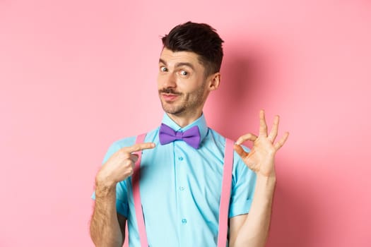 Image of young handsome guy in bow-tie and holiday outfit, pointing at himself and showing okay gesture, self-promoting on pink background.