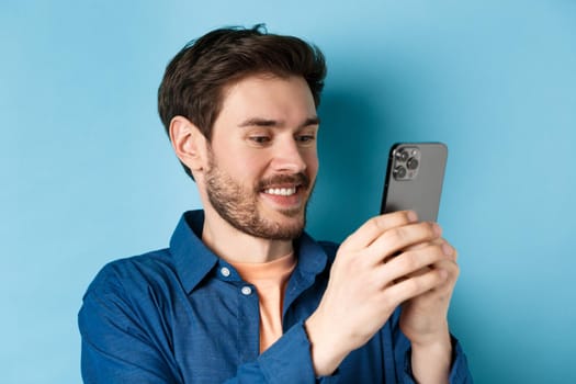 Close-up of handsome caucasian man writing message, reading mobile screen and smiling, standing on blue background.