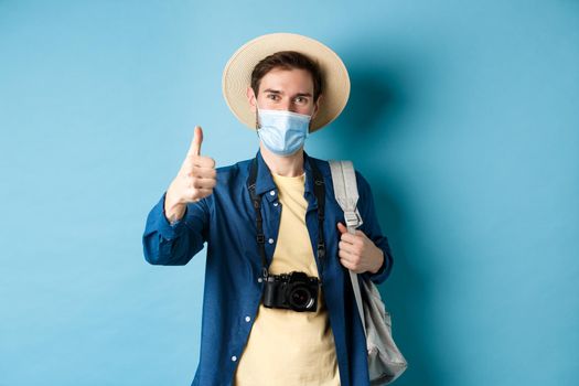 Covid-19 and summer holidays concept. Cheerful tourist in straw hat and medical mask praise and recommend good vacation hotel, showing thumb up, going on travel with backpack.