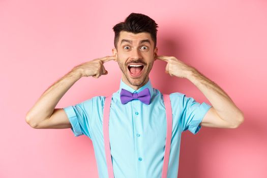 Childish funny guy shut ears and laughing at camera, refuse to listen, standing ignorant on pink background.