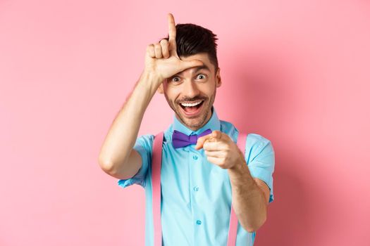 Image of funny guy pointing at you camera and showing loser sign, mocking person who lose, standing over pink background.