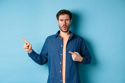 Shocked and offended young man frowning and looking hurt, pointing fingers left at empty space, complaining on something, standing on blue background.