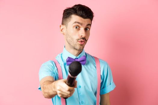 Handsome male show host stretch out hand with microphone, asking for interview or comments, standing on pink background.