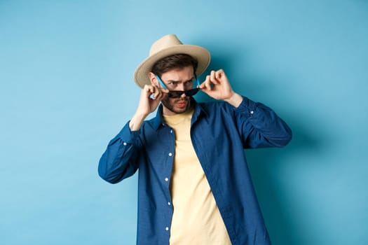 Portrait of guy on vacation staring aside with shocked and cringe face, take-off sunglasses and look at empty space on blue background.