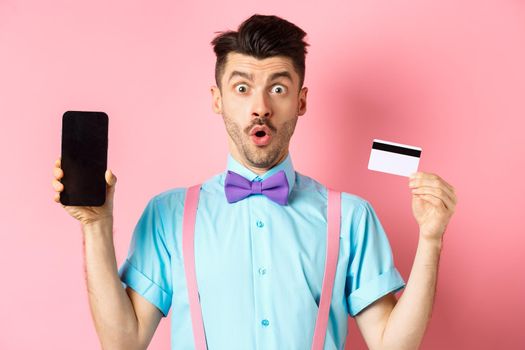 E-commerce and shopping concept. Surprised man showing blank smartphone screen with plastic credit card, standing amazed on pink background.