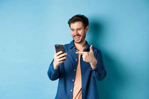 Image of handsome smiling man reading mobile screen and pointing at phone, checking out online promo or smartphone app, standing on blue background.